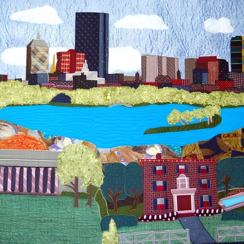 Pictorial art quilt by Rosie Rhine of Rhino Quilting portraying the Boston skyline, the MIT dome, and the commissioner's childhood home in Boston. Tall buildings in navy, brick red, tan, and plaid fabrics rise against a pale blue sky with white clouds. A band of leafy green trees lines the far side of a wide blue river, with a small tree-covered island in the water. At the left, a rust-orange dome and a columned building with striped fabric sit near the water’s edge. In the foreground, a red brick house with white-trimmed windows and a small cupola stands among green lawns, trees, and a walkway.