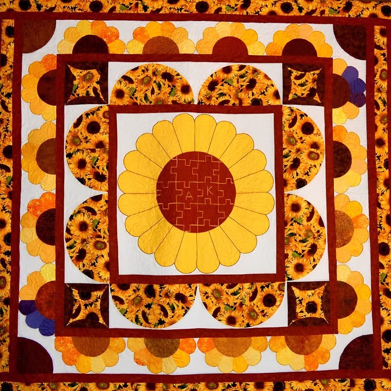 Sunflower-themed wedding quilt by Rosie Rhine of Rhino Quilting with a large sunflower at the center. Its petals are bright yellow, and its dark red center is pieced like a jigsaw puzzle with the couple’s initials stitched in. Rings of sunflower-print fabric and more flowers in yellow, orange, and deep red surround it, with scalloped petal shapes along the sides and corners on a white background. Dark red borders and a wide outer border of sunflower print frame the quilt.