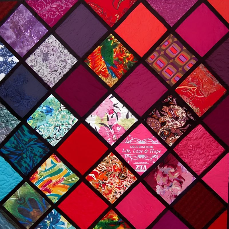 Close-up of a quilt by Rosie Rhine of Rhino Quilting with squares set on point and separated by black sashing. Solid squares in deep red, magenta, and plum alternate with squares of bright printed fabrics, including florals, a parrot, and abstract patterns. One pink square carries the printed words “Celebrating Life, Love & Hope.”