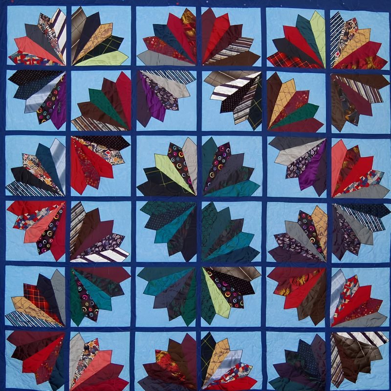 Bed-sized necktie quilt by Rosie Rhine of Rhino Quilting with a grid of fan-shaped blocks. Each fan is made of neckties in red, burgundy, green, teal, purple, black, and striped patterns, set on light blue squares. Dark navy sashing separates the blocks and frames the quilt.
