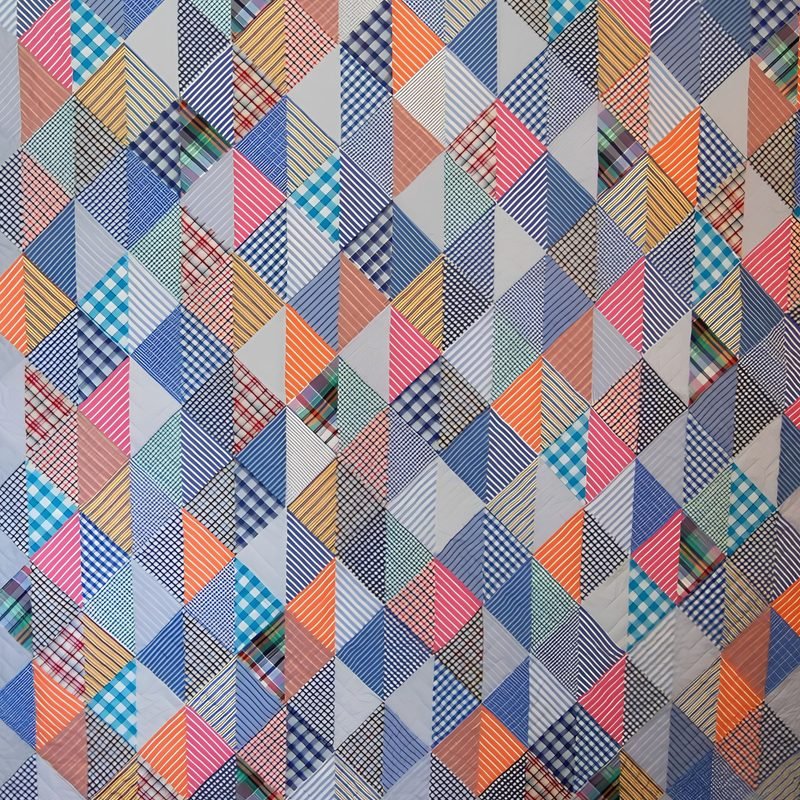 Close-up of a quilt by Rosie Rhine of Rhino Quilting made of small triangles in a repeating diamond pattern. The fabrics are mostly checks, stripes, and plaids in blue, gray, pink, orange, and teal, giving the surface a lively, scrappy look.