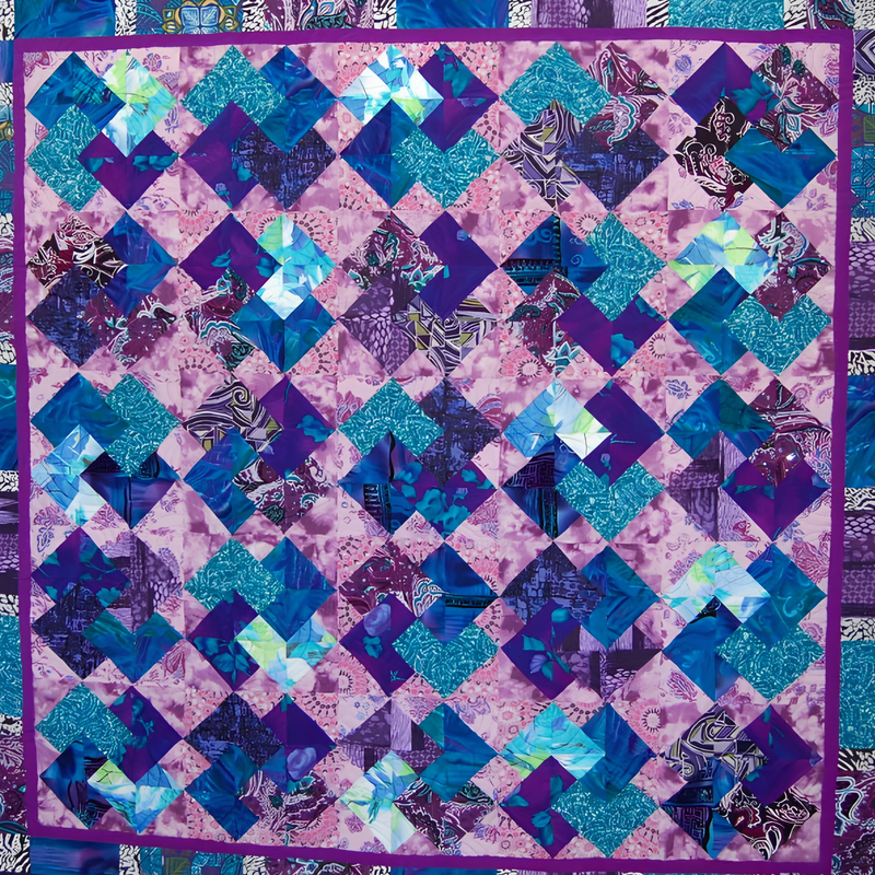 Quilt by Rosie Rhine of Rhino Quilting in the Card Trick block pattern, with overlapping squares that look like tucked playing cards. The cards are cut from fabrics in purple, deep blue, and teal, including glittery turquoise, marbled blue-green, and black and white paisley prints. They sit on a mottled lilac background, and a narrow purple border frames the quilt.