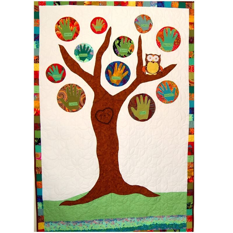 Appliqué quilt by Rosie Rhine of Rhino Quilting showing a large brown tree with a heart carved into its trunk, set on a cream background above a strip of green grass. Round fabric leaves in red, orange, green, and teal each hold a handprint, and a small owl sits on one of the branches. A border of multicolored fabric strips runs around the edge, and swirling quilting covers the background.