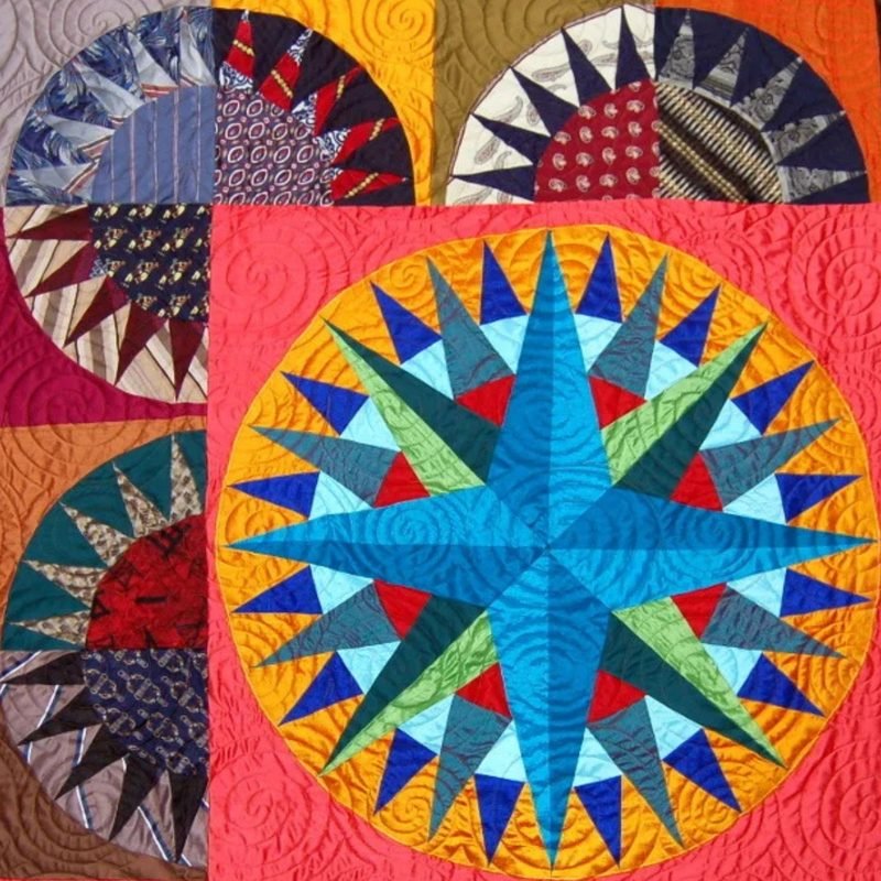 Detail shot of a quilt by Rosie Rhine of Rhino Quilting featuring a large compass rose in bright blue, with points in teal, green, red, and navy, set inside a gold ring on a coral-red background. Above and to the left, partial circular fan blocks are pieced from men’s shirt and necktie-style fabrics in navy, burgundy, gray, and teal, radiating like sunbursts. Swirling quilting lines are visible across the coral background.