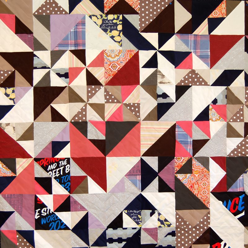 Quilt by Rosie Rhine of Rhino Quilting made of angular triangles in an abstract, Cubist-inspired arrangement. The triangles are cut from cream, brown, tan, gray, red, coral, and black fabrics, including polka dots, plaids, and paisley. Pieces of black and blue concert T-shirts with partial white and red lettering are mixed in. Fine quilting stitches run across the surface.