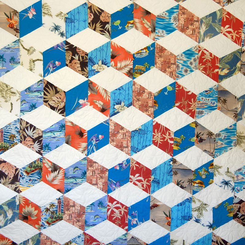 Close-up of a tumbling blocks quilt by Rosie Rhine of Rhino Quilting. Diamond shapes in cream, blue, and red tropical Hawaiian-style prints with palm trees and flowers are arranged to look like three-dimensional cubes. The cream diamonds carry fine quilting stitches.