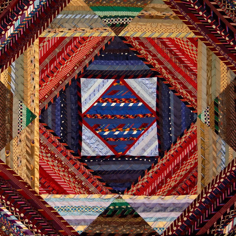 Necktie quilt by Rosie Rhine of Rhino Quilting in a concentric square-in-square design built from narrow tie strips. At the center, a small diamond of blue, red, and white striped ties is outlined in red. Rings of dark navy, red, and gold strips spread outward, ending in triangles at the edges. Most of the fabrics are striped or patterned ties in deep red, navy, gold, and brown.