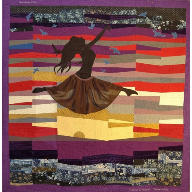 Pictorial bereavement quilt of a dancer’s silhouette leaping across a sunset sky, her sheer brown skirt flaring out. The sky is pieced from horizontal strips of purple, red, gray, gold and cream fabric, with blue birds flying around her and a setting sun below. Dark blue and purple floral fabrics form the ground, framed by a purple border.