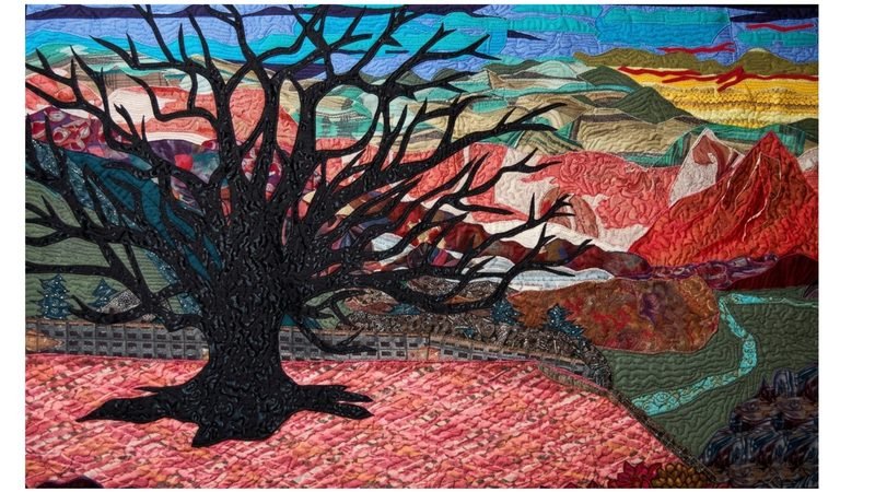 Art quilt by Rosie Rhine of Rhino Quilting depicting a dramatic mountain landscape. In the foreground, a massive black tree with a thick trunk and a web of bare branches fills the left side, its roots spreading across a field of textured coral and pink fabric. A low gray stone wall runs behind the tree, and small evergreen trees dot the hillsides. To the right, a teal and green river winds through the valley. Beyond it, overlapping ridges in red, orange, rust, and olive lead up to a bright red peak. The sky shifts from blue and turquoise at the top to yellow and gold near the horizon, with streaks of red cloud. Dense, swirling quilting stitches add texture across the whole piece.
