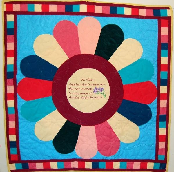 Small quilt by Rosie Rhine of Rhino Quilting with a large flower of rounded petals in pink, cream, black, navy, teal, and red around a dark red ring. The cream center holds a printed message with a small purple flower: “For Violet, Grandma’s love is always near. This quilt was made in loving memory of Grandma Zylpha Newcome.” The flower sits on a bright blue background with a striped multicolored border.